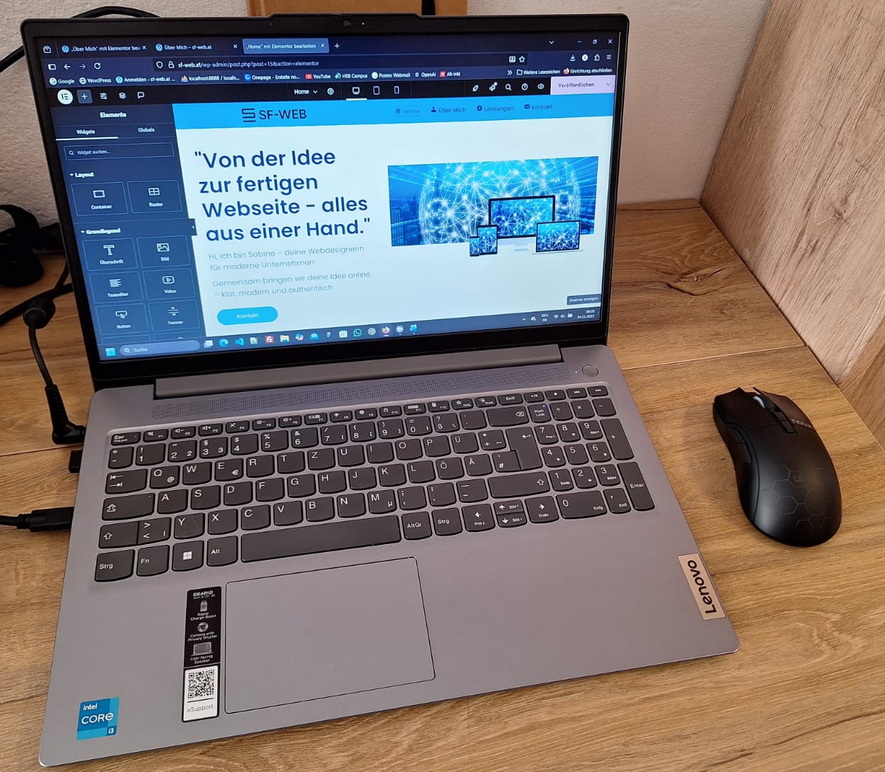 Arbeitsplatz mit einem geöffneten Lenovo Notebook mit Intel Core i3 Aufkleber, auf dem das Backend einer Webseite mit dem Slogan „Von der Idee zur fertigen Webseite – alles aus einer Hand.“ und dem SF-WEB Logo in einem Elementor Builder angezeigt wird. Rechts liegt eine schwarze Maus auf einem Holztisch.