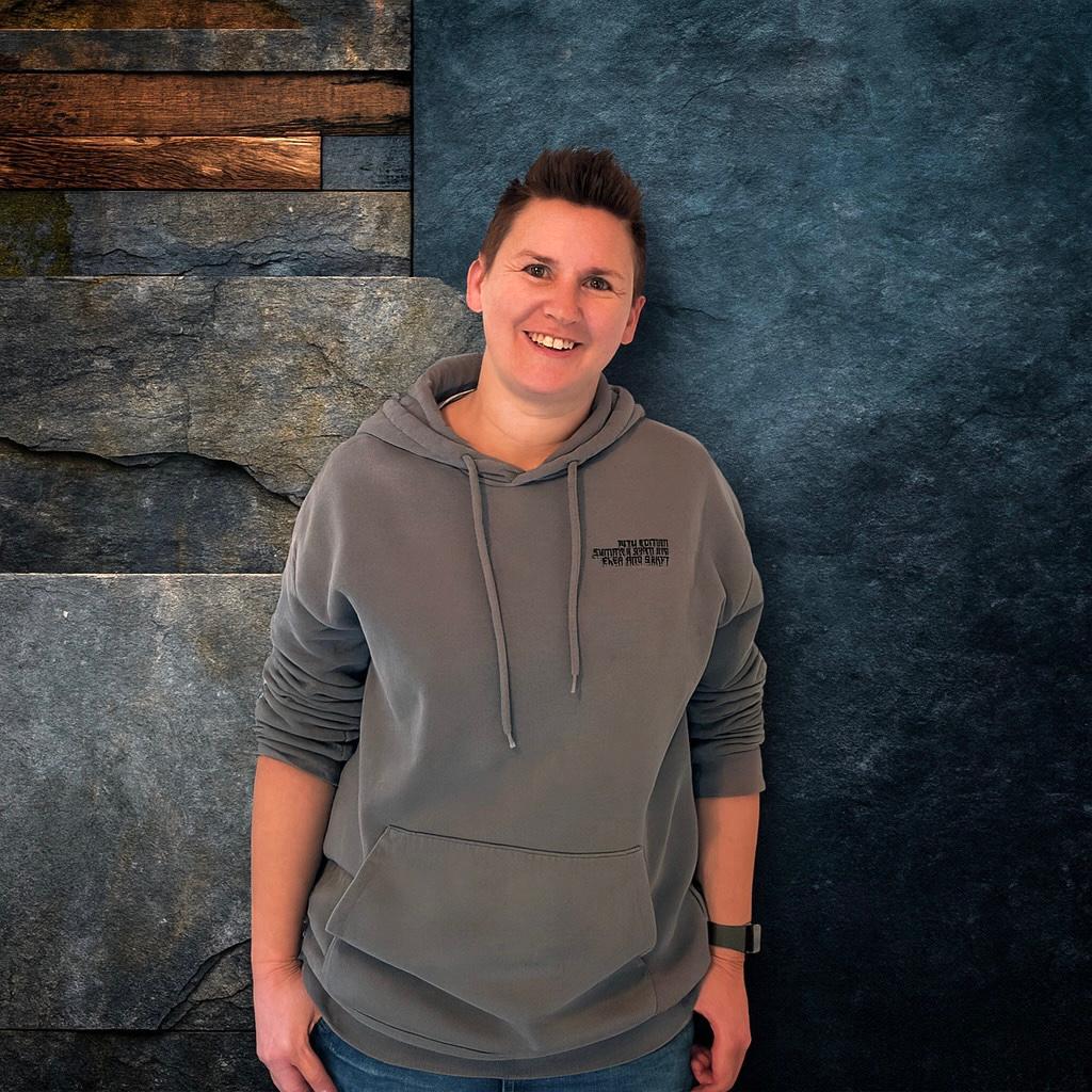 Kurzhaarige Person in einem grauen Hoodie und Jeans l&auml;chelt vor einer dunkelblauen und grauen Steinwand.