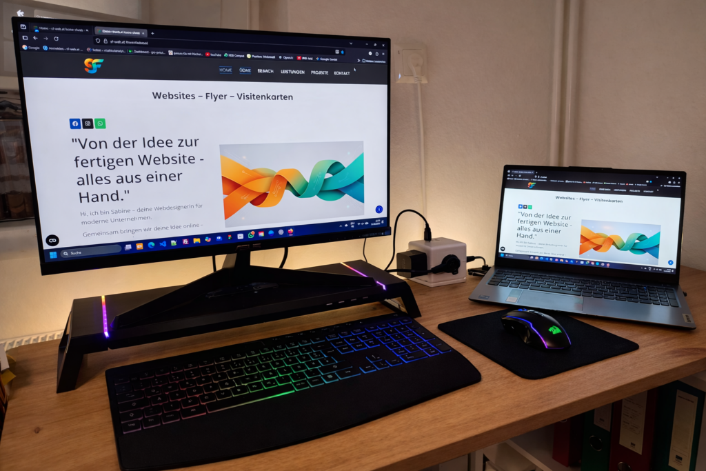 Professioneller Arbeitsplatz mit einem großen Monitor und einem Laptop. Auf beiden Bildschirmen ist die Webdesign-Portfolio-Website von Sabine (SF) zu sehen, mit dem Logo und dem Text: 'Websites – Flyer – Visitenkarten' und der Überschrift: 'Von der Idee zur fertigen Website – alles aus einer Hand.' Der Schreibtisch aus Holz zeigt eine ergonomische Tastatur und Maus mit farbiger Beleuchtung.